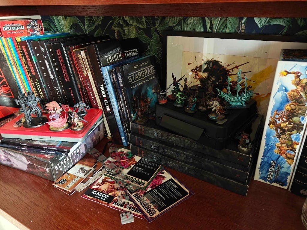 Wooden shelf with skirmish wargame rulebooks, boxed sets, painted miniatures in a carry case, and tabletop game cards.